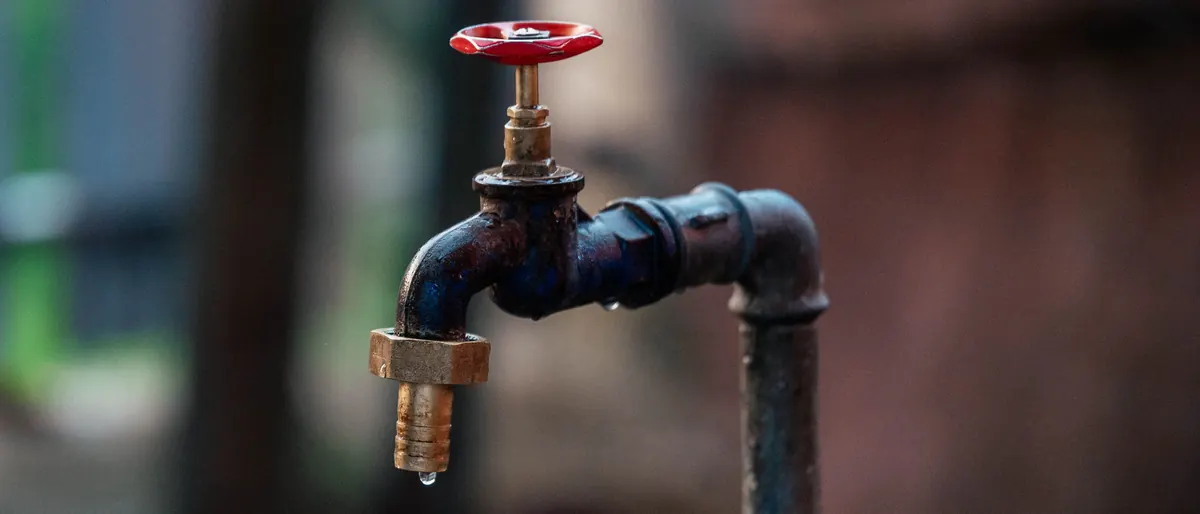 Alter Außenwasserhahn mit rotem Griff, ein Tropfen hängt am Auslauf